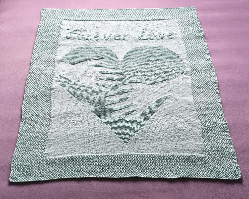Light green and white knitted baby blanket showing two hands forming a heart and 'Forever Love' stitched above, with a textured border.