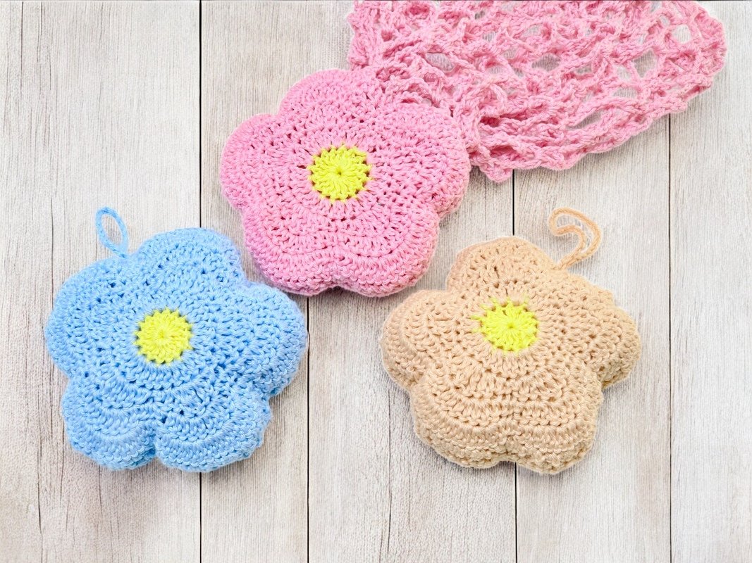 Three crocheted flower appliqués in pink, blue, and beige with yellow centers on a light wooden surface beside a pink openwork crochet fabric.
