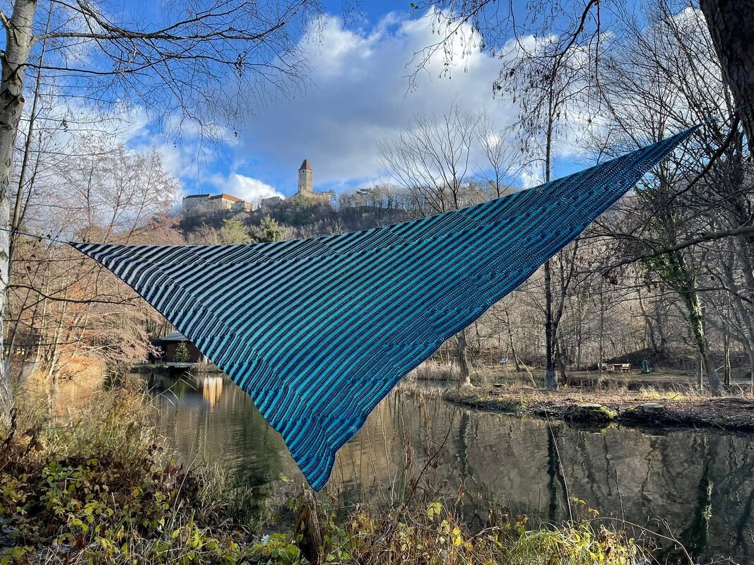 Blau-türkis gestreiftes Dreieckstuch aus Häkelarbeit hängt zwischen Bäumen über einem See, im Hintergrund eine Burg auf einem Hügel.