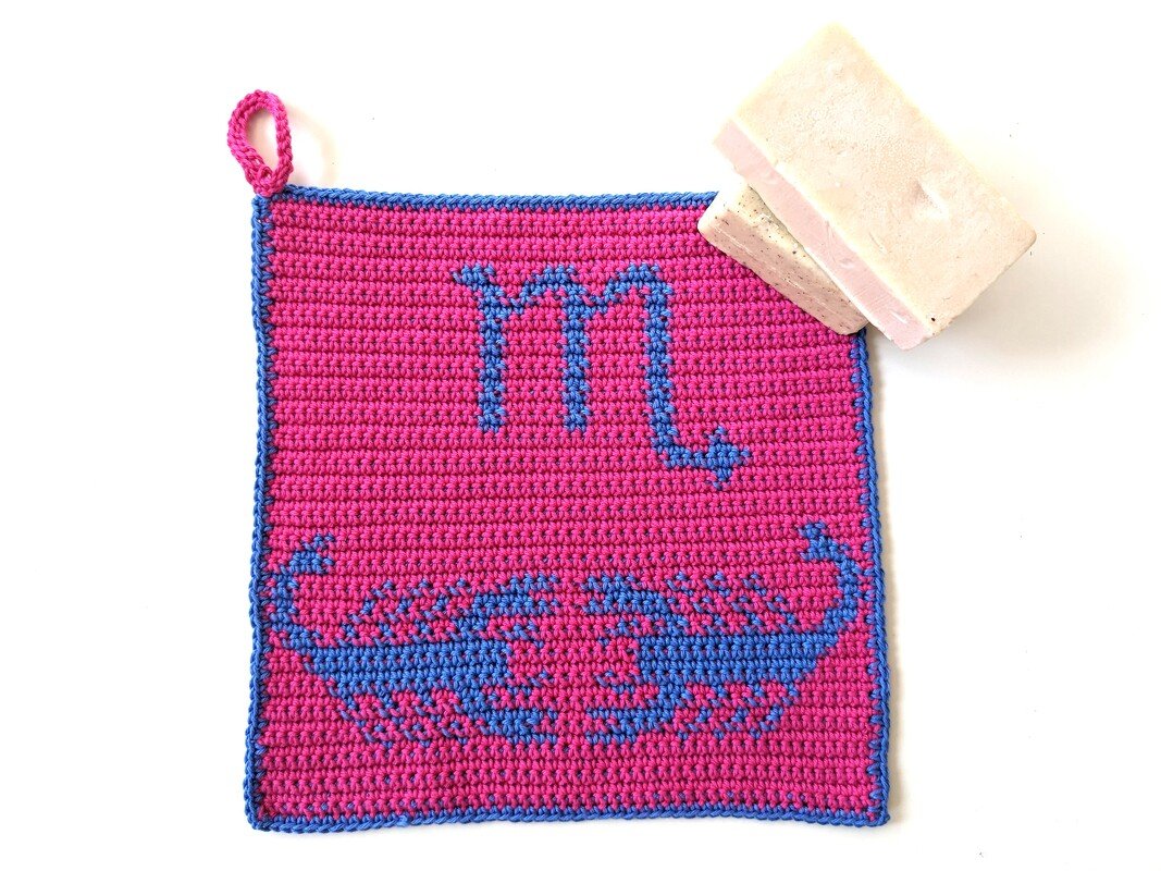 Quadratischer gehäkelter Topflappen in Pink mit blauem Skorpion-Motiv und Aufhängeschlaufe auf weißem Hintergrund
