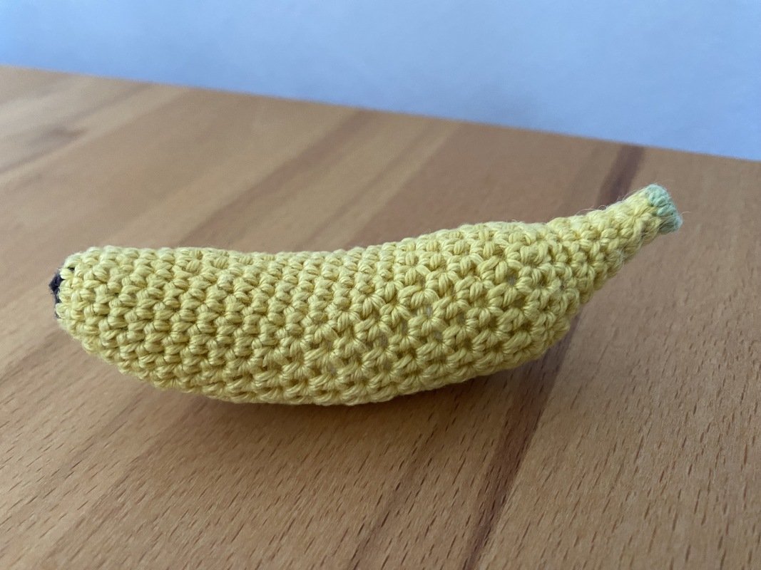 Gelbe gehäkelte Banane mit grünem Stiel und dunkler Spitze auf hellem Holztisch.