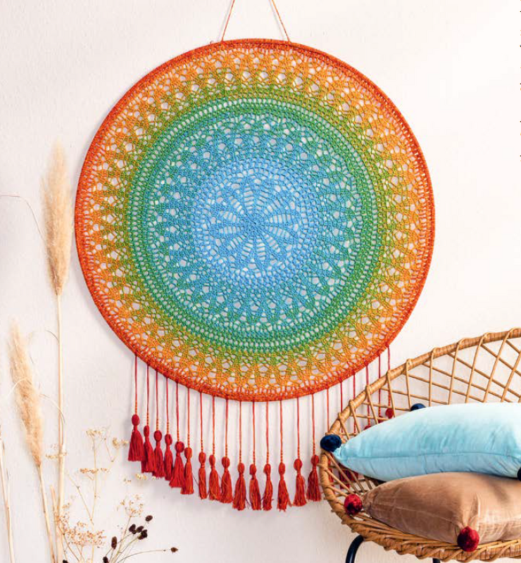 Runder gehäkelter Mandala-Wandbehang in Regenbogenfarben mit roten Quasten an weißer Wand