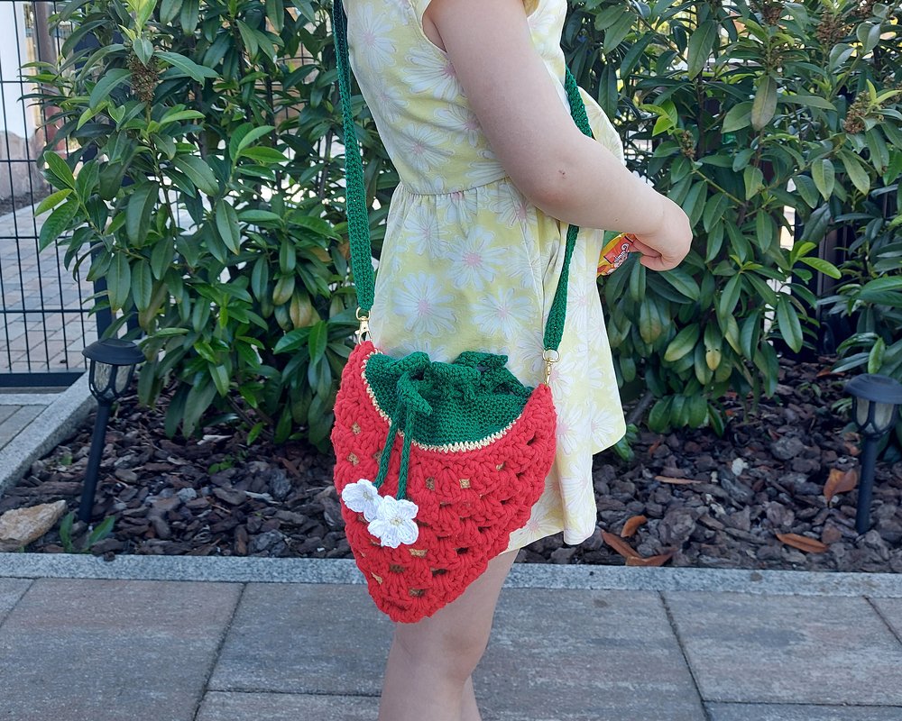 Kind trägt gehäkelte Erdbeer-Umhängetasche: rot mit grünem Zugband, grünem Schultergurt, goldfarbenen Karabinern und weißer Häkelblüte