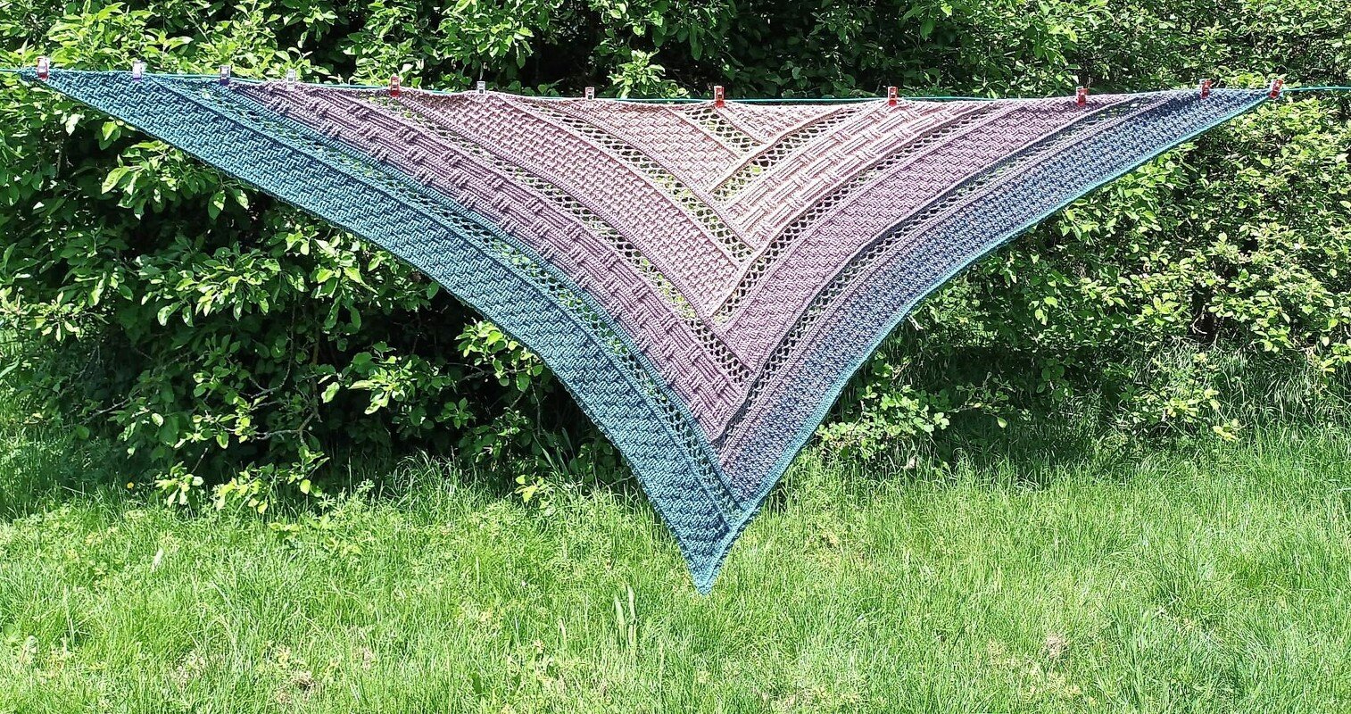 Blocked triangle crochet shawl with teal-to-lilac gradient hung on a line against green foliage