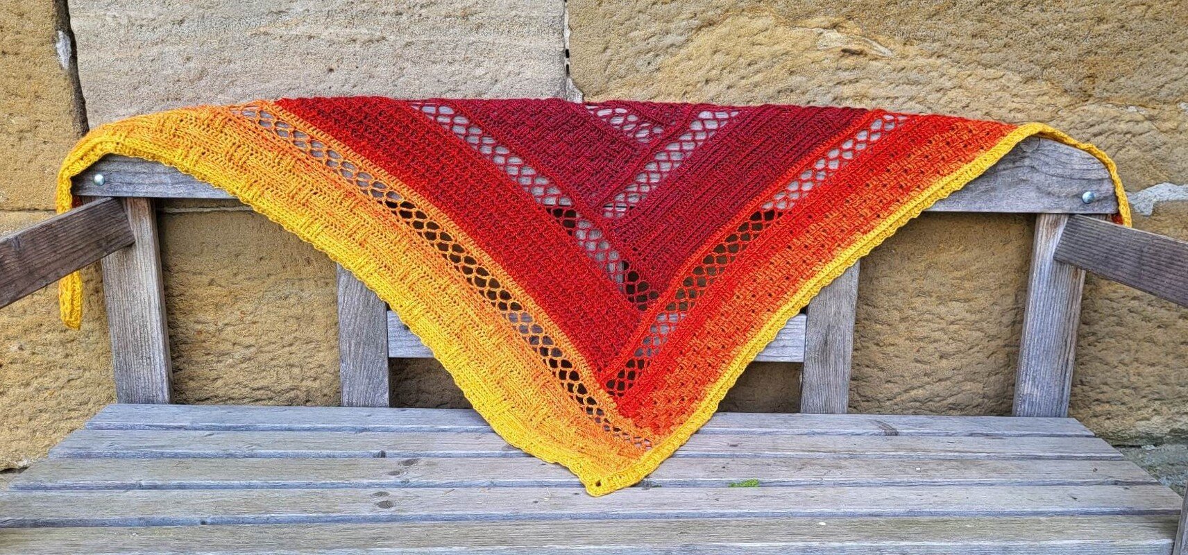 Triangular crochet shawl in yellow, orange and red with lace stripes draped over a wooden bench