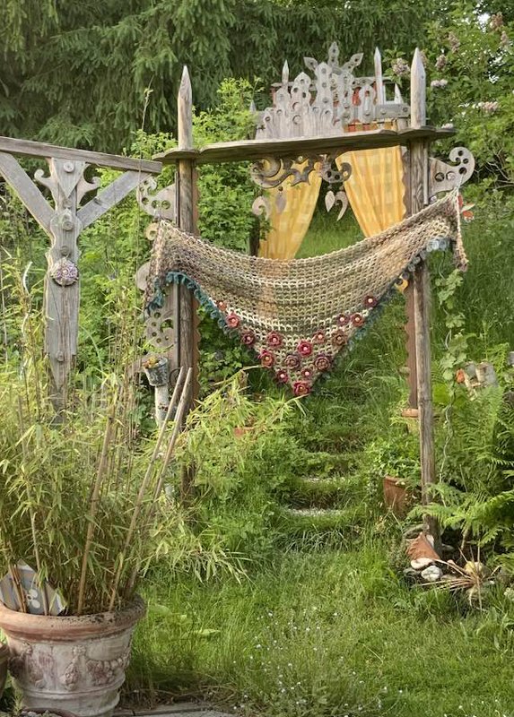 Beigefarbenes, handgehäkeltes Dreieckstuch mit roter Blumenborte an einem geschnitzten Holzbogen im Garten