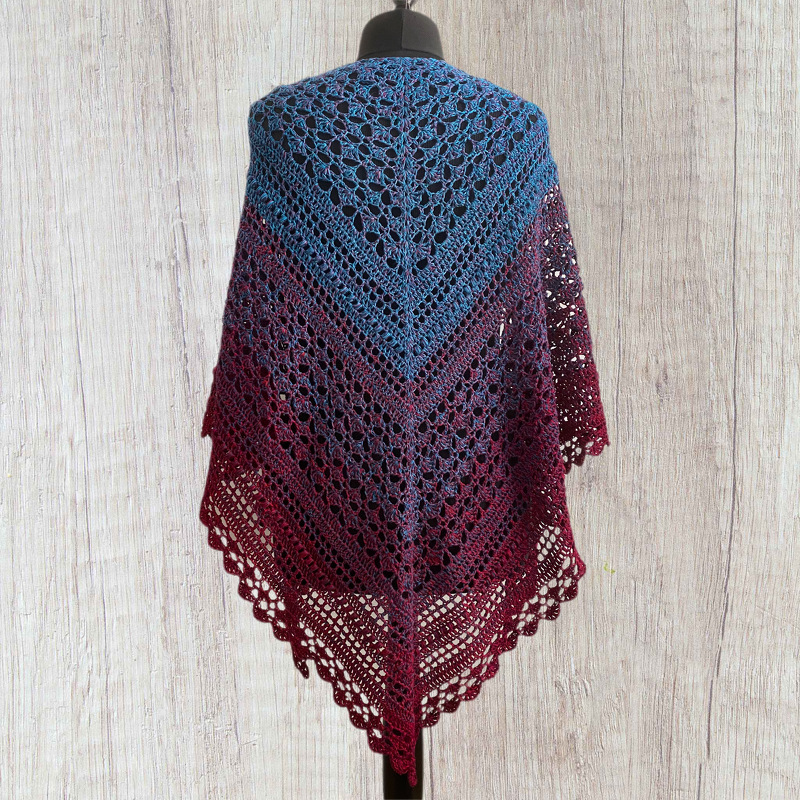 Crocheted triangular shawl with a blue-to-burgundy ombre, draped on a dress form against a wooden backdrop.