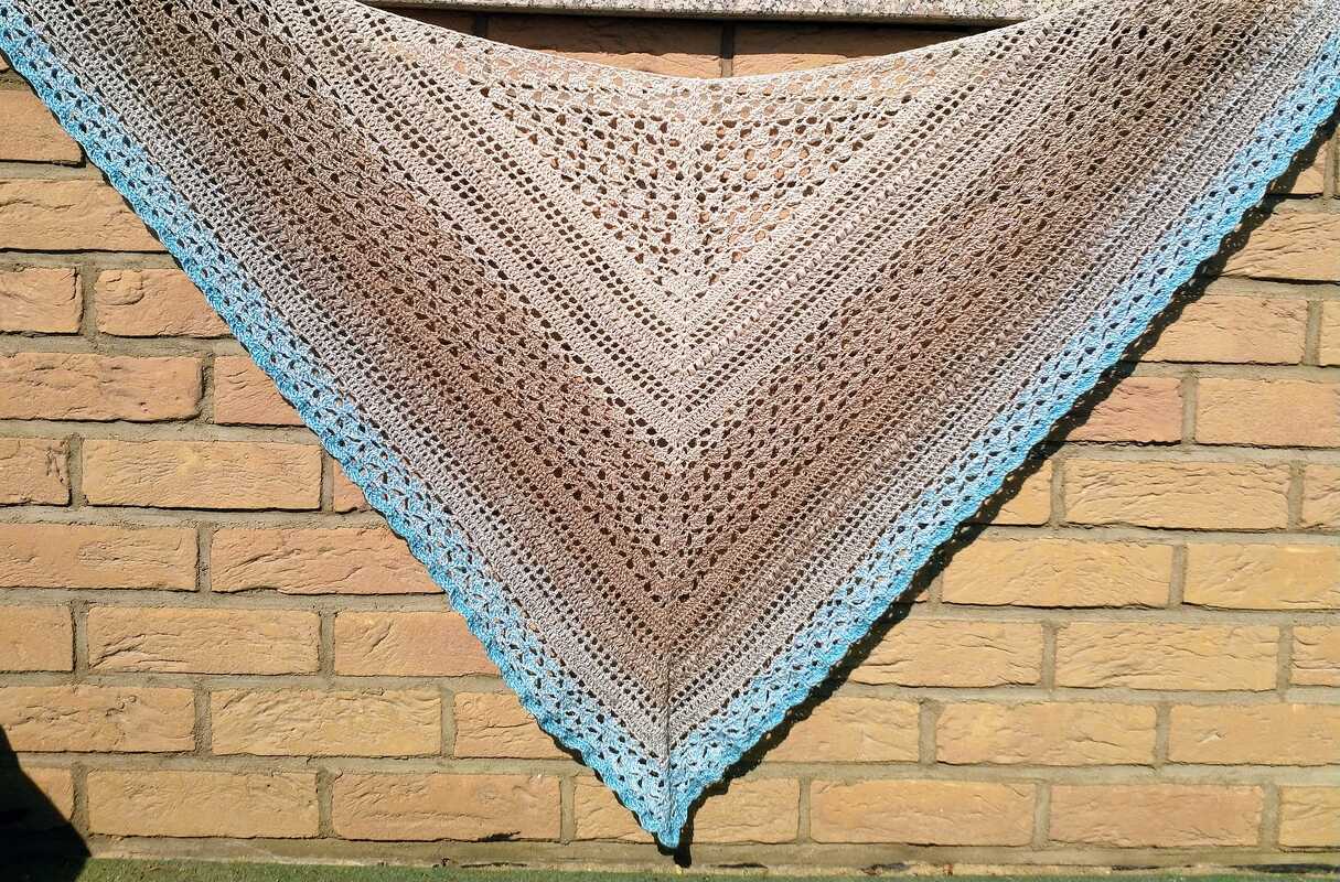 Ombré crochet triangle shawl with lacy openwork and bright blue scalloped border hanging on a brick wall.