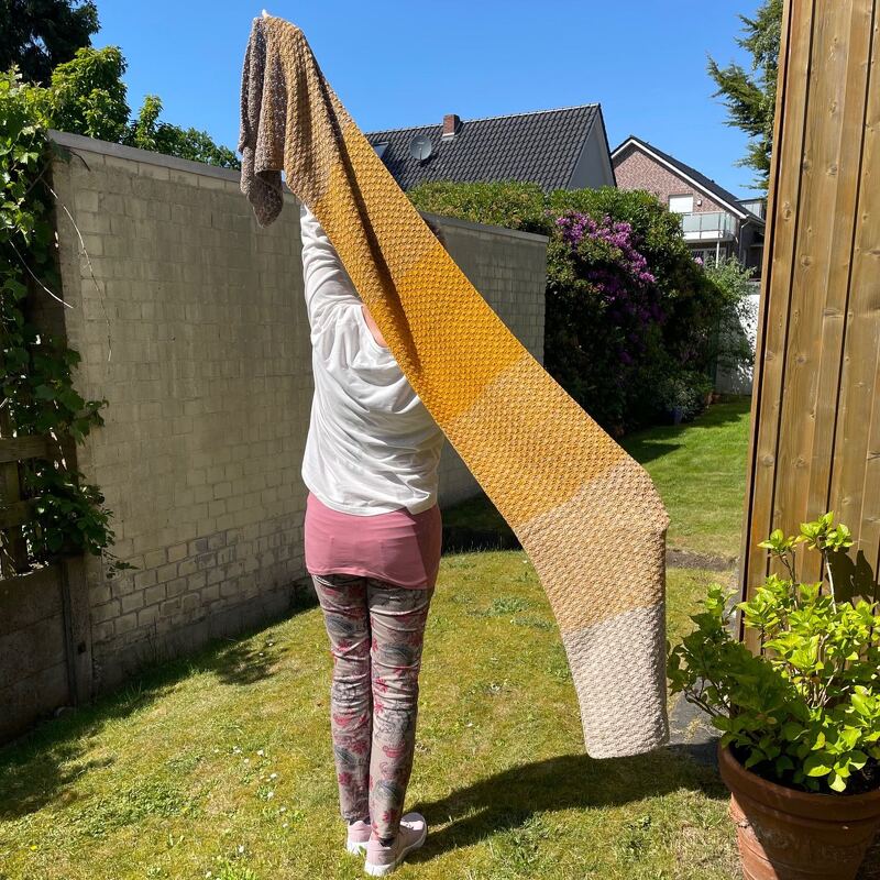 Person von hinten hält einen langen, gelb-beige verlaufenden Häkelschal senkrecht vor einem Gartenhintergrund.