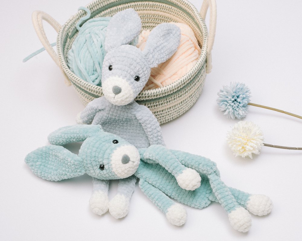 Two soft crochet bunny loveys in pale blue and light gray next to a basket of yarn and pom-pom flowers.