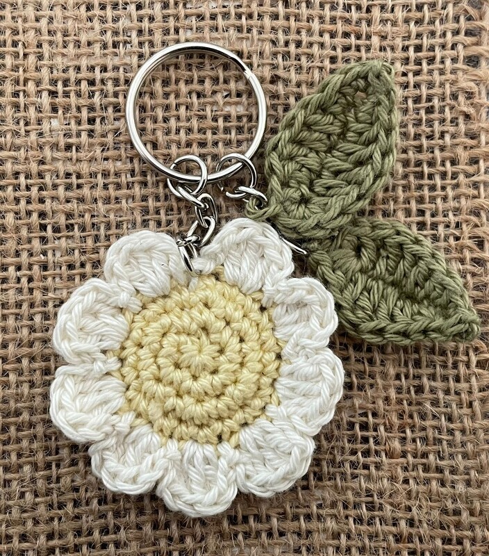 Nahaufnahme eines gehäkelten Gänseblümchen-Schlüsselanhängers mit cremeweißen Blütenblättern, hellgelber Mitte und zwei grünen Blättern an einem Metallring auf Jute.