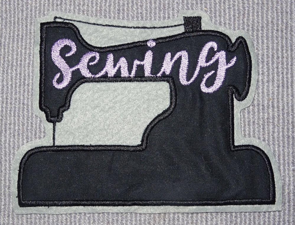 Gesticktes Nähmaschinenmotiv in Schwarz mit lila ‚Sewing‘-Schriftzug auf hellgrauem Grund