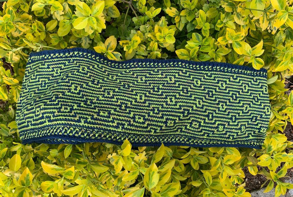 Gehäkelter Loop in Dunkelblau und Limettengrün mit geometrischem Muster auf gelben Blättern