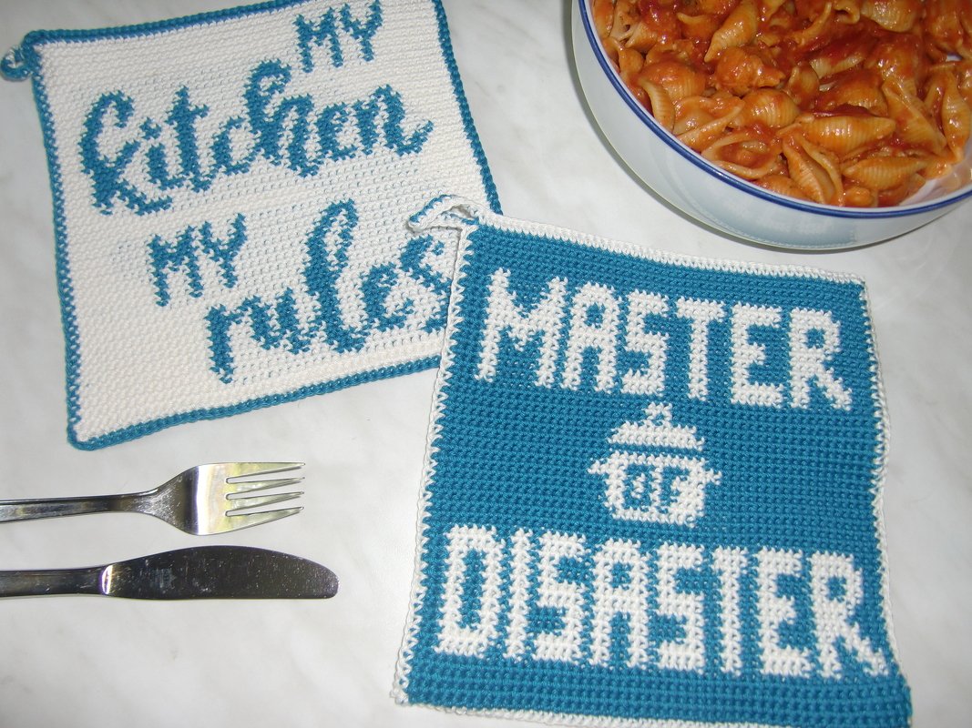Two square teal and white crochet potholders with text 'My kitchen my rules' and 'MASTER DISASTER' beside a bowl of pasta and cutlery