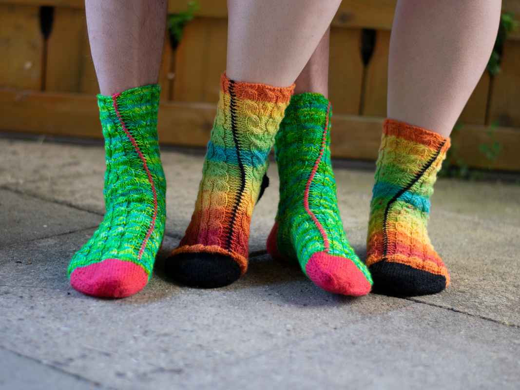 Two people wearing colorful hand-knitted cable socks in gradient yarn with contrasting heels and toes