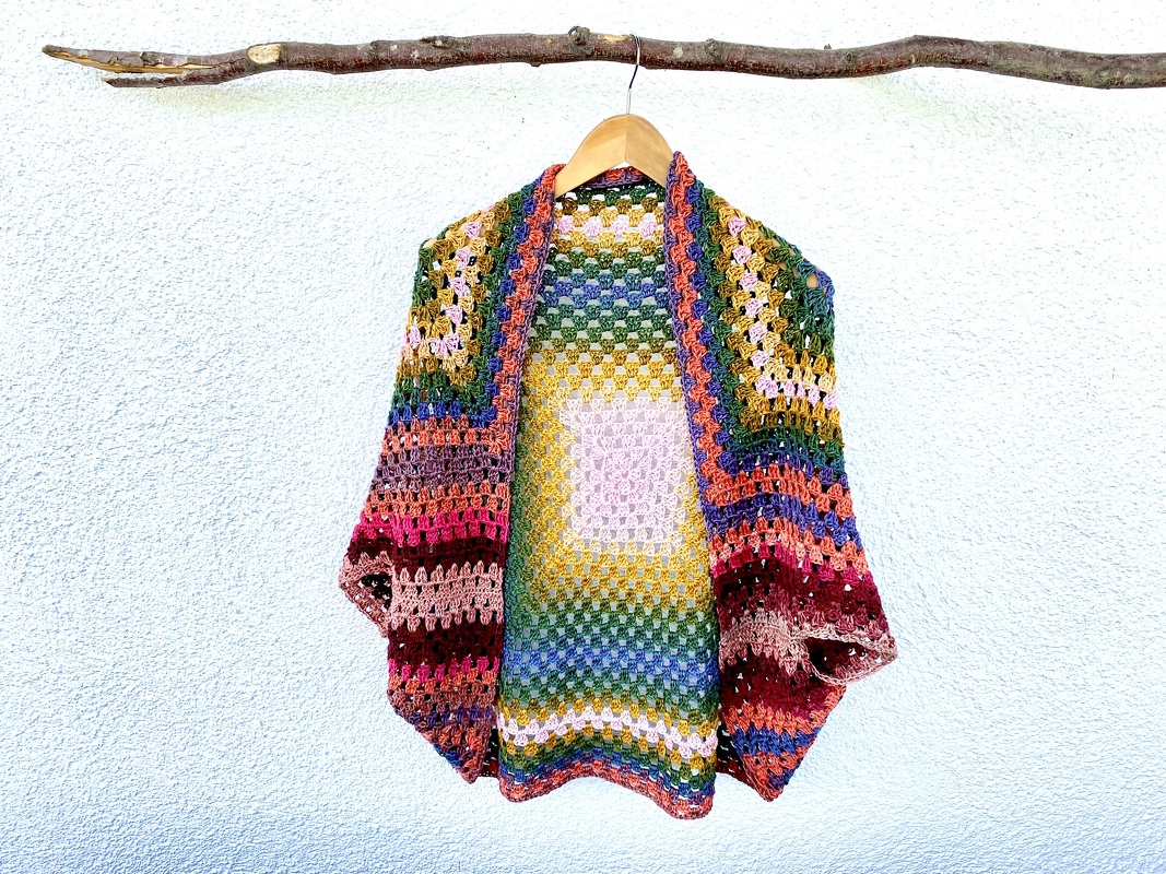 Bunter gehäkelter Seelenwärmer mit Granny‑Muster an einem Holzbügel vor heller Wand