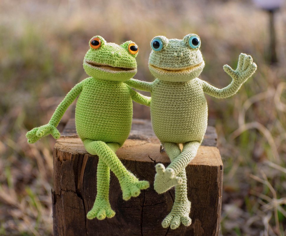 Two green crocheted amigurumi frogs with orange and blue eyes sitting on a wooden stump.