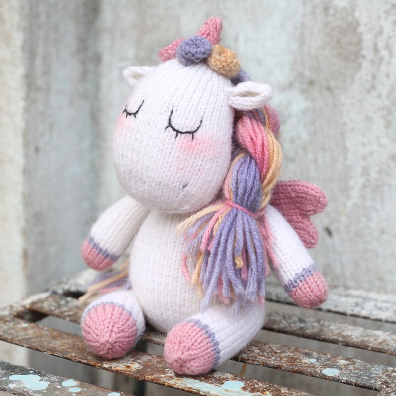 White knitted unicorn amigurumi with braided pastel mane, pink wings, and embroidered closed eyes