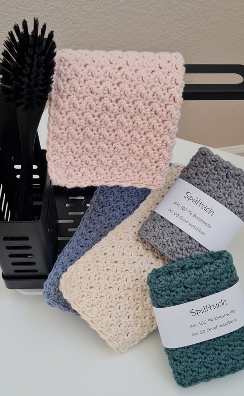 Gehäkelte Spültücher aus Baumwolle in rosa, blau, beige, grau und grün, teilweise gerollt mit Etikett neben Küchenbürste und Halter.