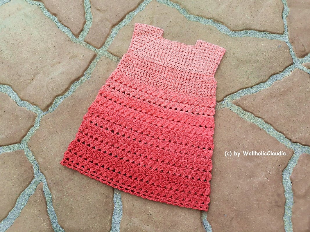 Ärmelloses gehäkeltes Babykleid in Rosa bis Korall, flach auf Steinboden gelegt, mit Loch- und Muschelmuster