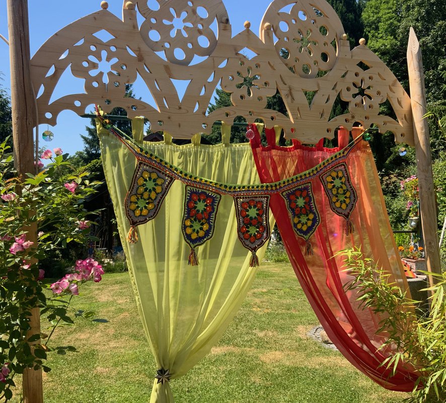 Gehäkelte Banner-Girlande mit bunten Mustern und Quasten an einer geschnitzten Holzpergola neben grünen und roten Vorhängen