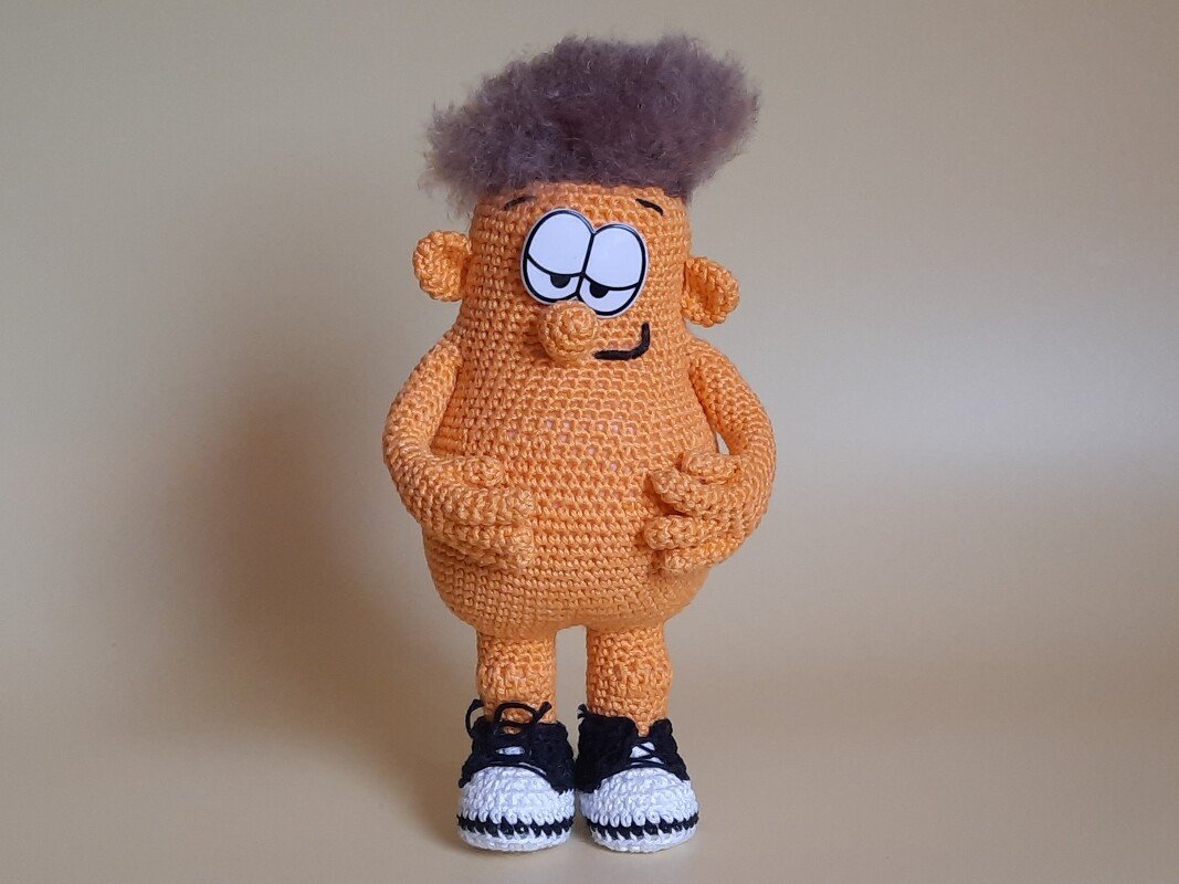 Orangefarbene Amigurumi-Figur mit grauen Wuschelhaaren und gehäkelten Turnschuhen