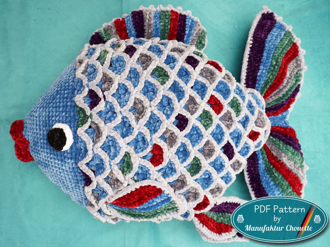 Gehäkeltes Fischkissen in Blau mit weißem Schuppenraster und mehrfarbigen Schuppen auf türkisfarbenem Hintergrund.