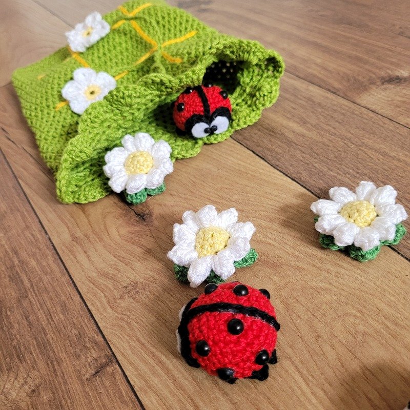 Zwei gehäkelte rote Marienkäfer und mehrere weiße Gänseblümchen um eine grüne Häkeltasche auf einem Holzboden