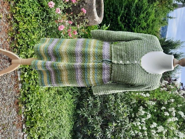 Mannequin mit grünem langärmeligen Häkeloberteil, durchgehender Knopfleiste und mehrfarbig gestreiftem Rock im Garten