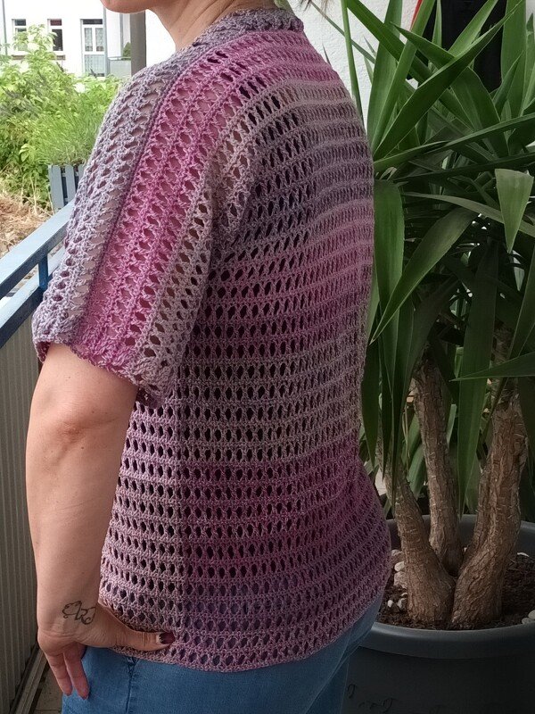 Person trägt kurzärmeliges, gehäkeltes Oberteil in Flieder- und Beerentönen mit Lochmuster auf einem Balkon.