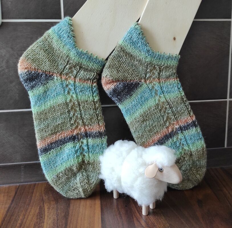 Paar gestrickter knöchelanger Socken in grünen, blauen und pfirsichfarbenen Streifen mit seitlichem Ajourmuster auf Holz-Sockenspannern, davor kleine Schäfchenfigur