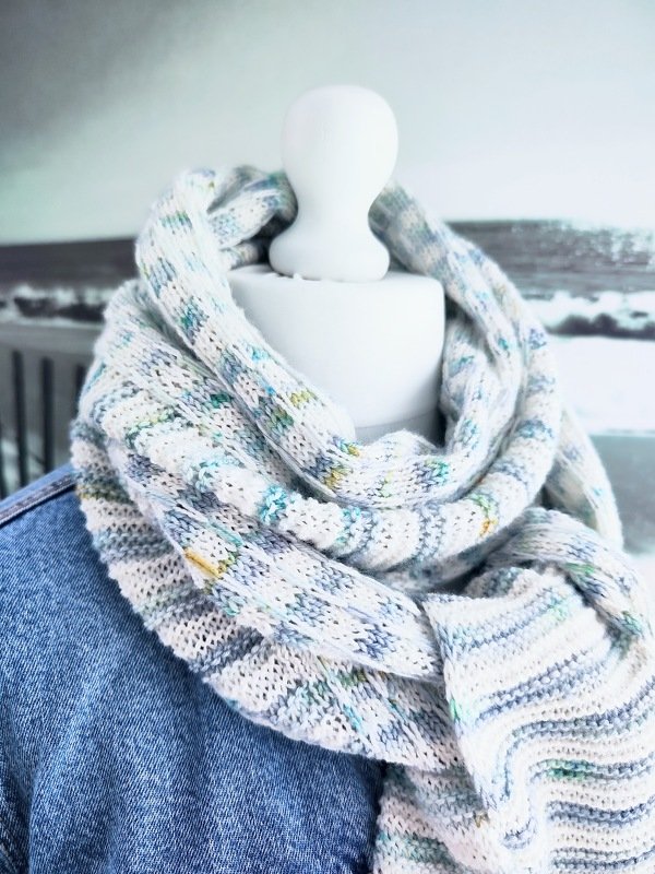 Heller gestrickter Loop-Schal mit blauen, grünen und gelben Sprenkeln, an einer Schaufensterpuppe über einer Jeansjacke drapiert.