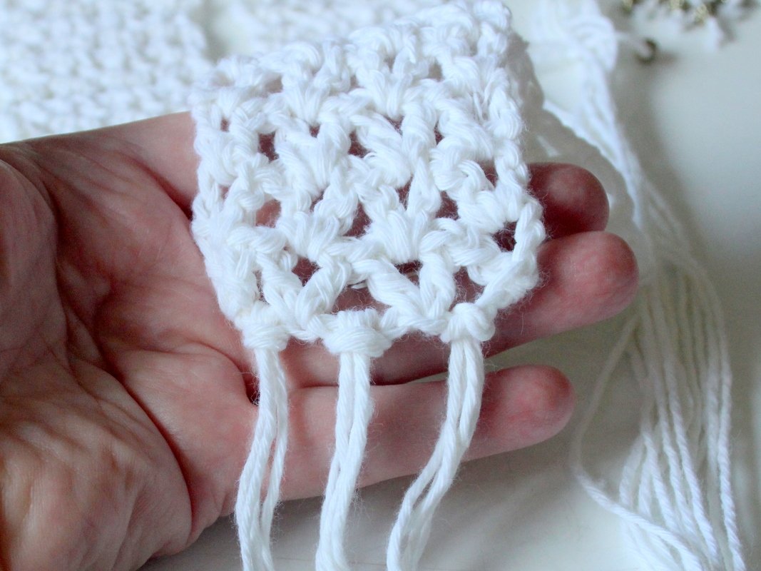 Hand hält ein weißes, gehäkeltes Muster mit offenen Maschen und kurzen Fransen