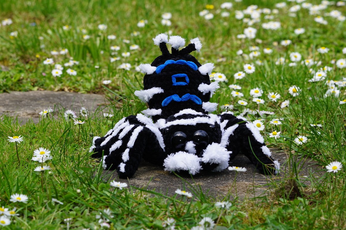 Gehäkelte Amigurumi-Spinne in Schwarz-Weiß mit blauen Akzenten auf einem Stein im Gras zwischen Gänseblümchen