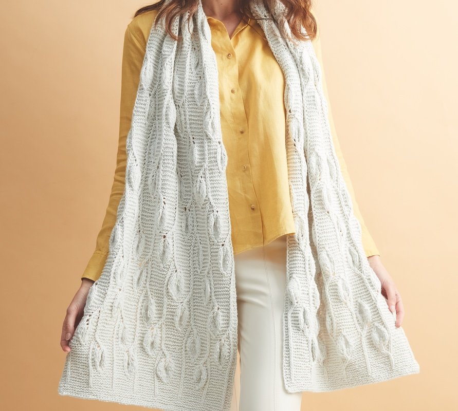 Person trägt langen cremeweißen Schal mit Blatt-Zopfmuster über gelber Bluse