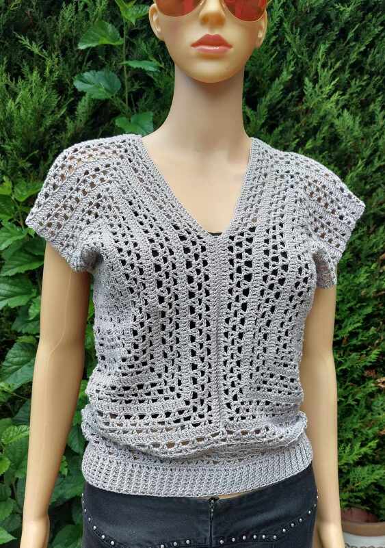 Grau gehäkeltes Kurzarm-Top mit V‑Ausschnitt an einer Schaufensterpuppe vor grünem Hintergrund.