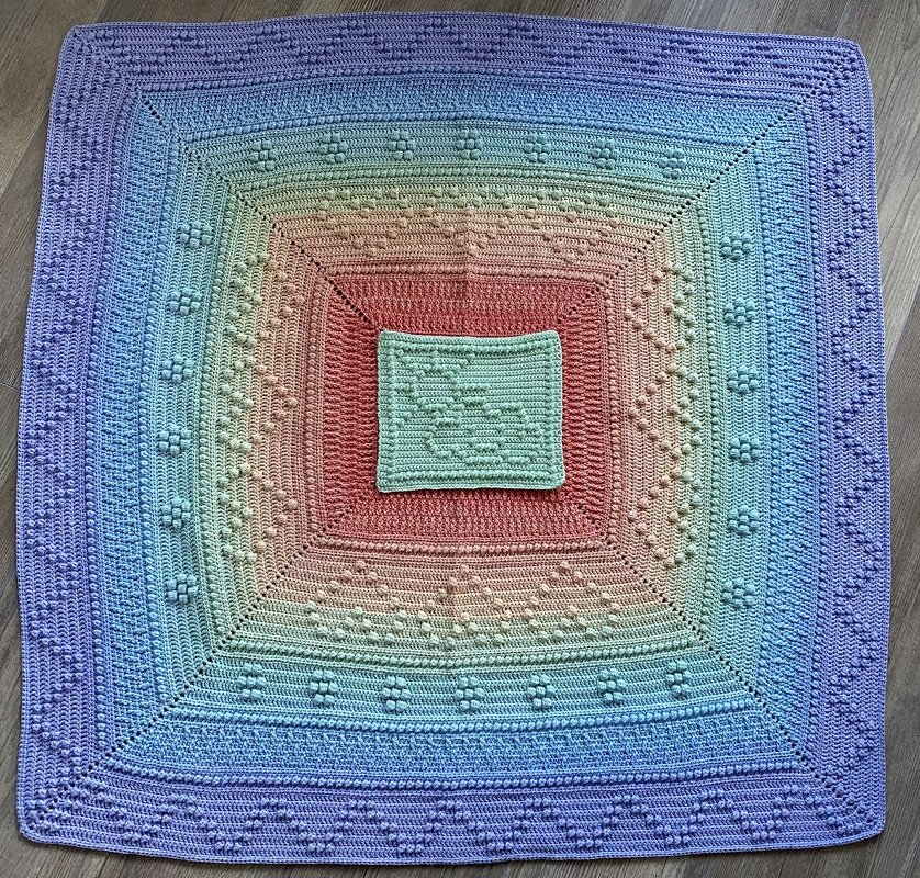 Quadratische gehäkelte Babydecke in pastelligen Regenbogenfarben mit erhabenen Bobbelmustern und gezacktem Rand, flach ausgelegt