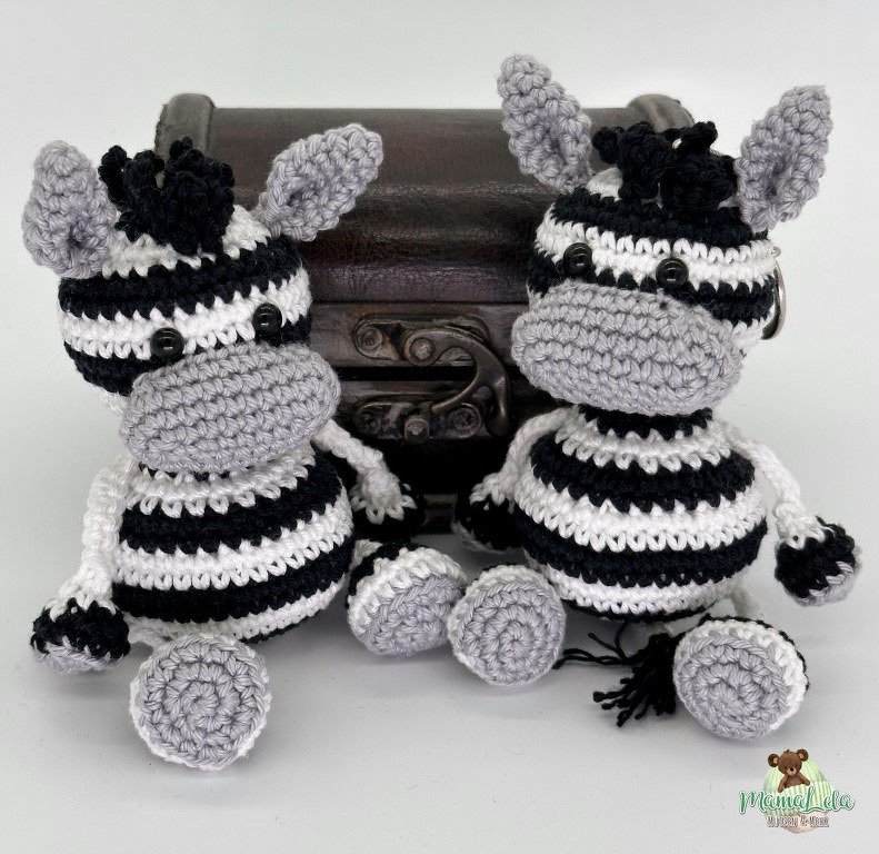 Zwei gehäkelte Zebra‑Amigurumi mit schwarzen und weißen Streifen, grauen Mäulern und Knopfaugen vor einer kleinen Truhe