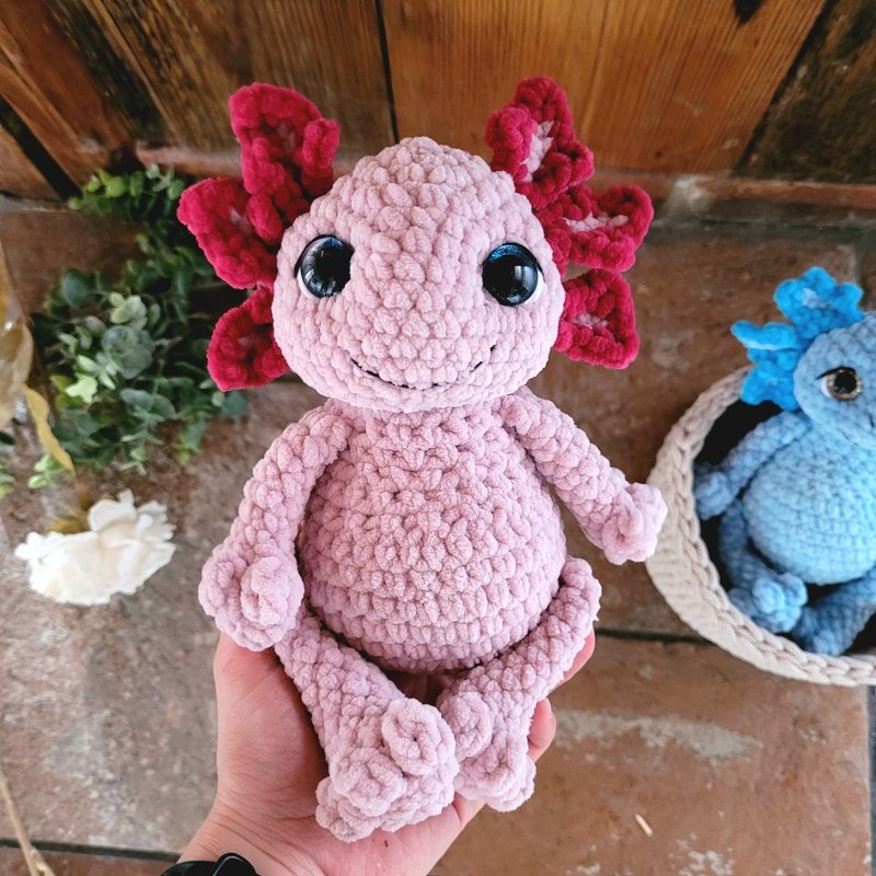 Hand hält gehäkeltes rosa Axolotl mit roten Kiemen und glänzenden Augen, im Hintergrund ein blaues Axolotl.