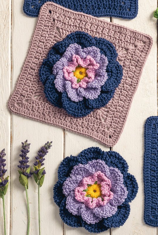 Zwei handgehäkelte Blumen, eine auf einem Granny Square, die andere separat, mit Lavendelzweigen auf hellem Holz.