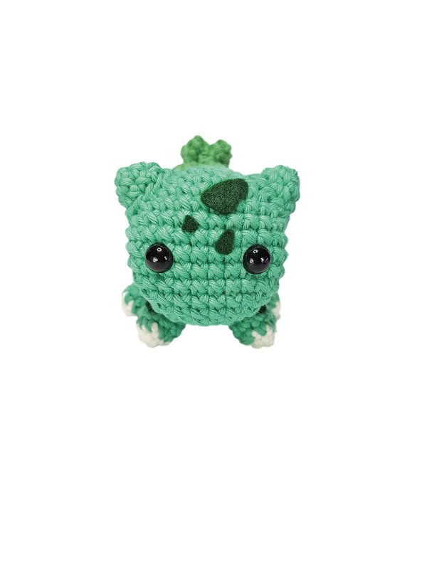 Grünes handgehäkeltes Amigurumi mit schwarzen Sicherheitsaugen, Blattapplikationen und weißen Pfoten vor weißem Hintergrund
