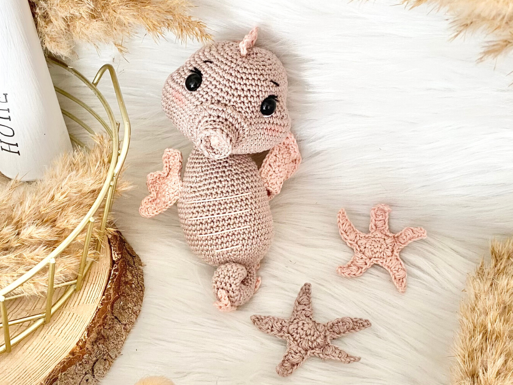 Gehäkeltes Amigurumi-Seepferdchen in Rosé mit zwei gehäkelten Seesternen auf hellem Untergrund