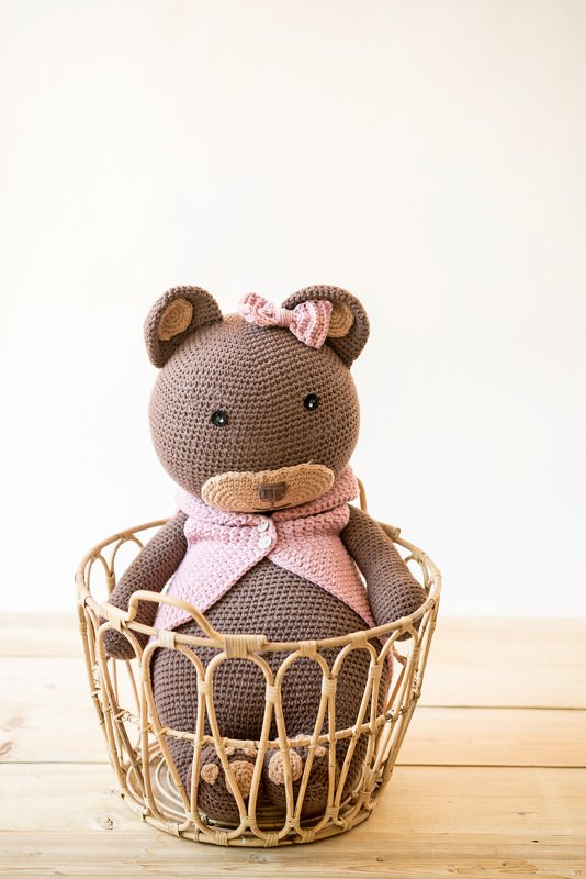 Brauner gehäkelter Teddybär mit rosa Weste und Schleife im Weidenkorb auf hellem Holz.