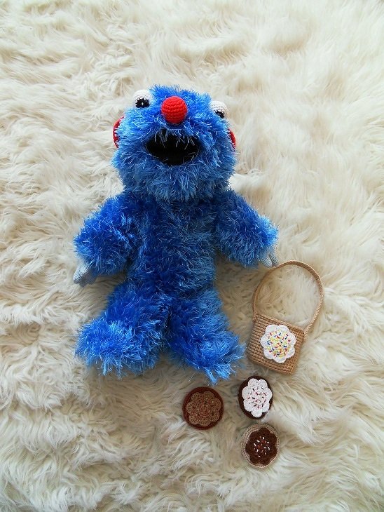 Blaues flauschiges Amigurumi mit roter Nase, kleinem Häkelkörbchen und runden Häkeldekorationen auf hellem Fell