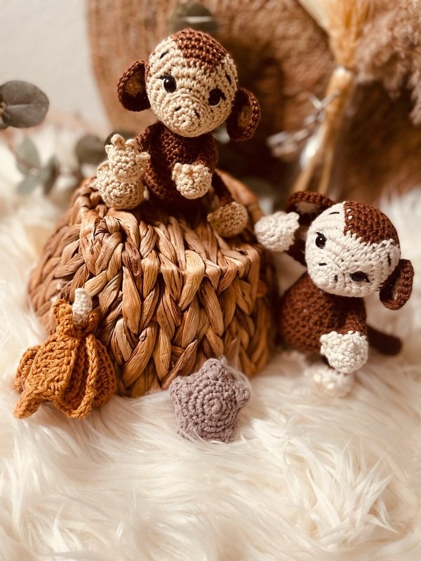 Zwei braun-beige gehäkelte Amigurumi-Affen auf einem geflochtenen Korb mit kleinen gehäkelten Accessoires auf flauschigem Untergrund.