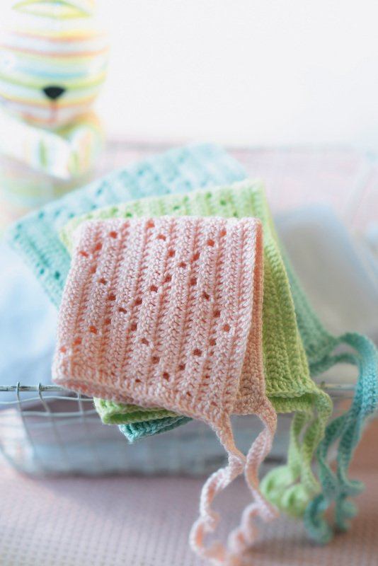 Stapel von drei gehäkelten Babymützen in Pastellrosa, Hellgrün und Mintblau mit Lochmuster und Bindebändern im Drahtkorb.