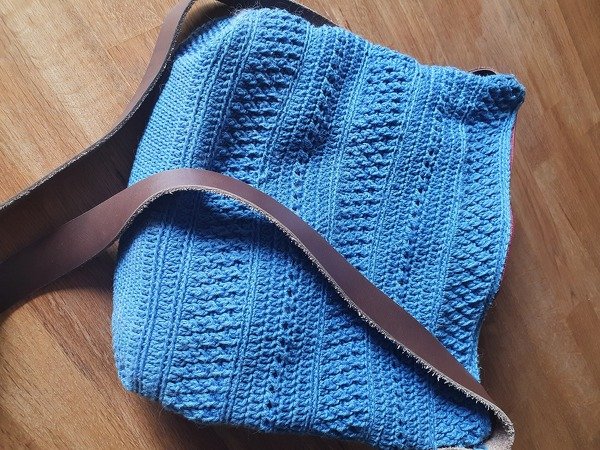 Blaue gehäkelte Umhängetasche mit braunem Lederriemen auf Holzboden