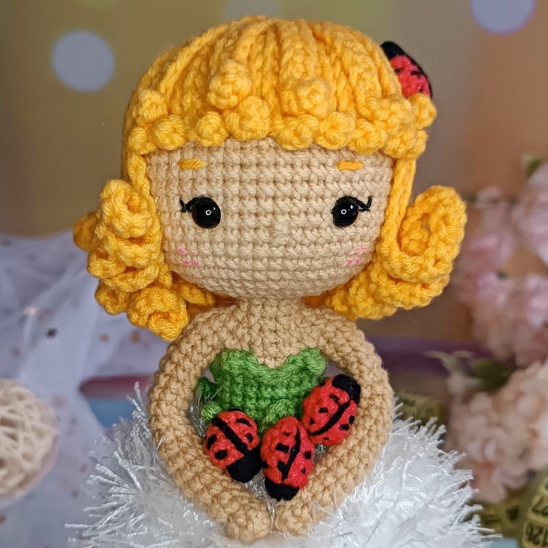 Crocheted amigurumi doll with yellow curly hair, black eyes, pink cheeks, holding three ladybugs and a small green leaf.