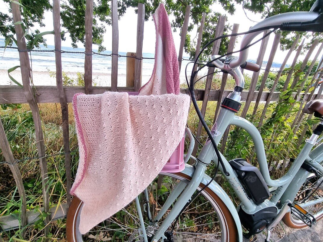 Zartrosa gestricktes Dreieckstuch am Fahrradkorb mit Strand im Hintergrund
