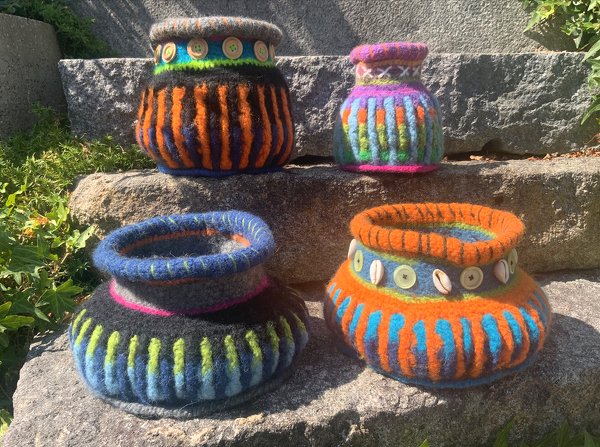 Vier bunte gehäkelte, gefilzte Körbe mit Rippenmuster und Knopf-/Muschelverzierung auf steinernen Stufen im Freien